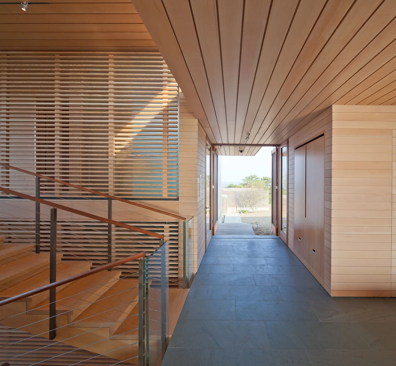 The hallway of Island Residence features wood paneling on walls and ceiling with a stone tile floor and a wooden staircase with glass railing.