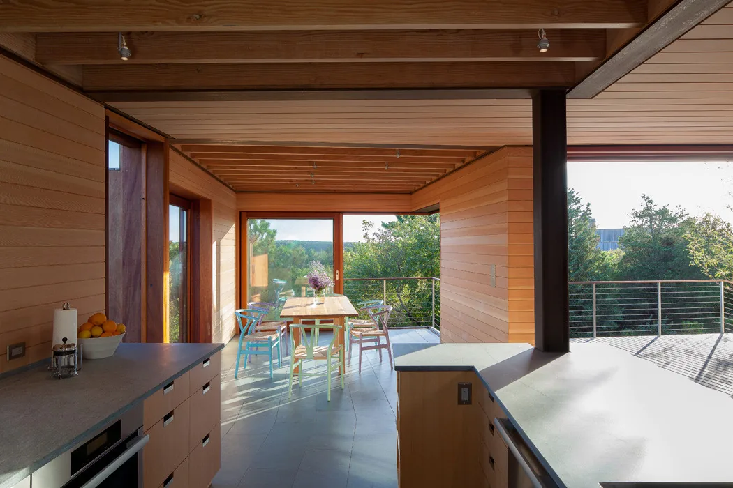 The Island Residence kitchen and dining area features wood paneling and large sliding glass doors to an outdoor deck with steel railing.