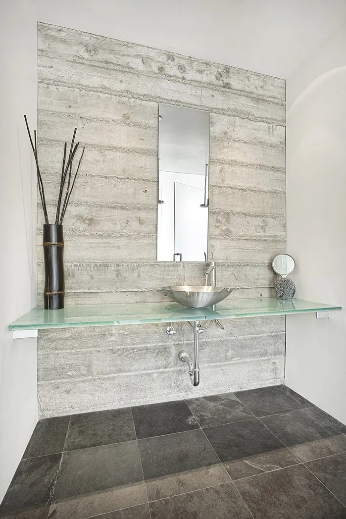 Bathroom features a glass countertop with a metal vessel sink and concrete textured wall.