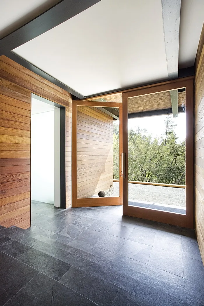 Entry hall of Lovell Residence features a wood-paneled wall and a large glass pivot door opening to the outdoors.