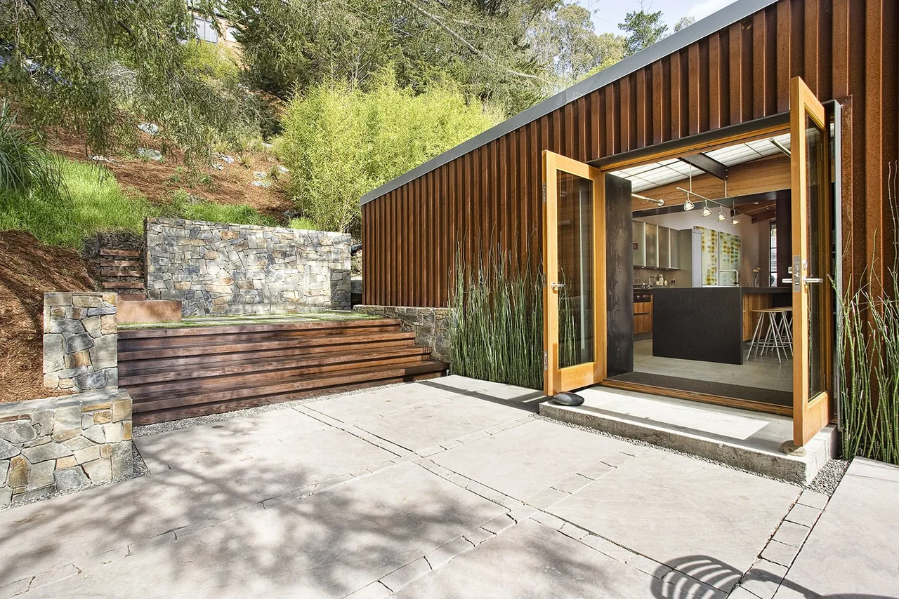 The Lovell Residence features rusted steel siding, stone landscaping walls, and open wooden doors connecting the kitchen to the outdoor patio.