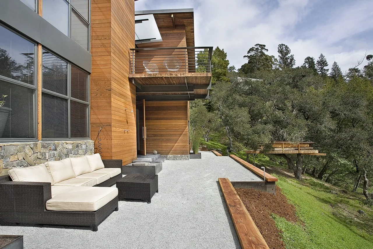 Modern exterior of Lovell Residence featuring a wood and stone facade with outdoor seating and a balcony overseeing a grassy slope.