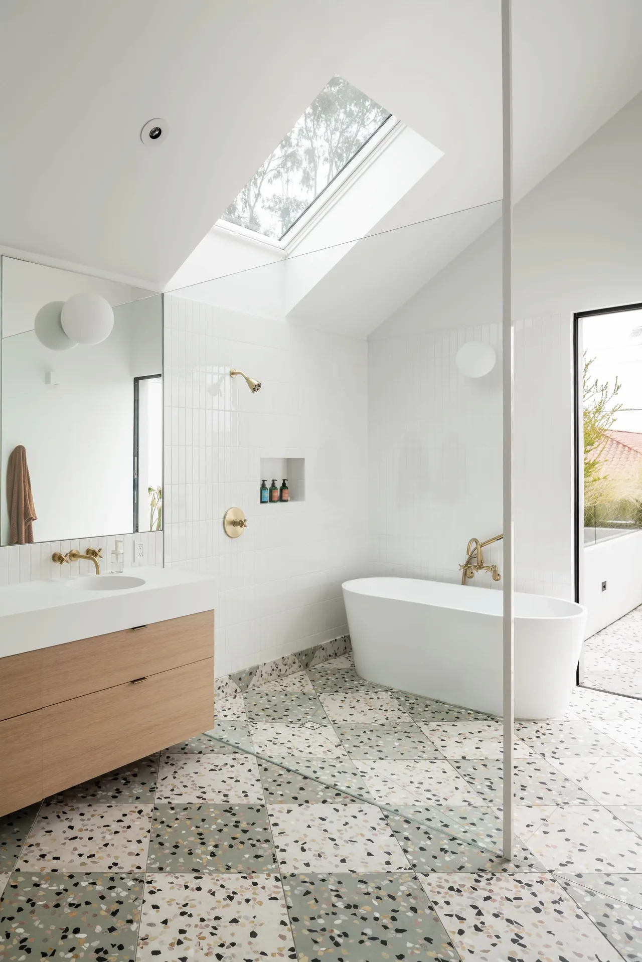 Modern bathroom featuring terrazzo floor tiles, a freestanding tub, skylight, and gold fixtures at Oak Avenue House.