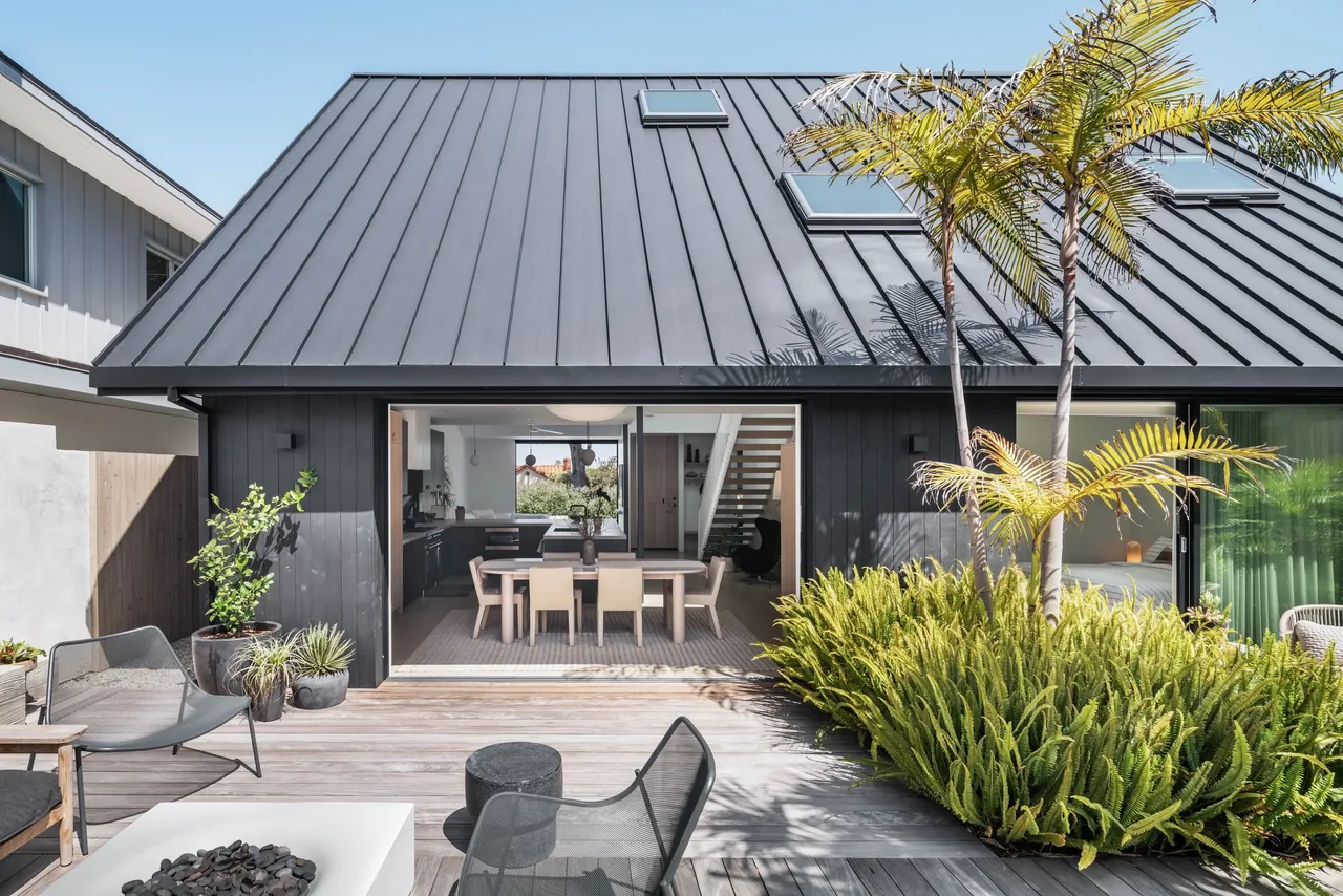 The Curtis Avenue House features a standing seam metal roof and black wood siding, opening through sliding glass doors onto a landscaped wooden deck patio.