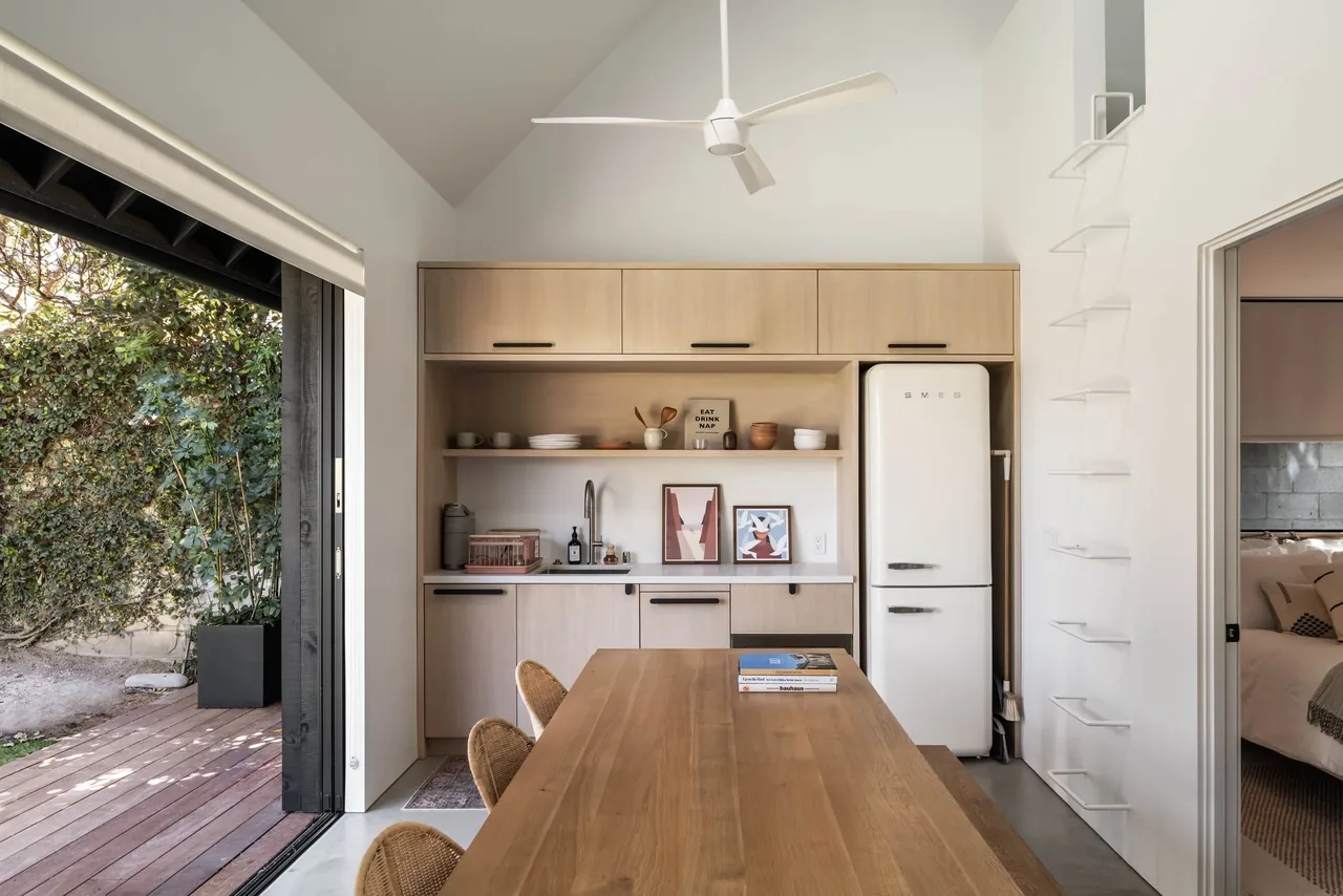 Light wood kitchen cabinetry with open shelving, white SMEG fridge, and a wooden dining table adjacent to sliding door to outside space.
