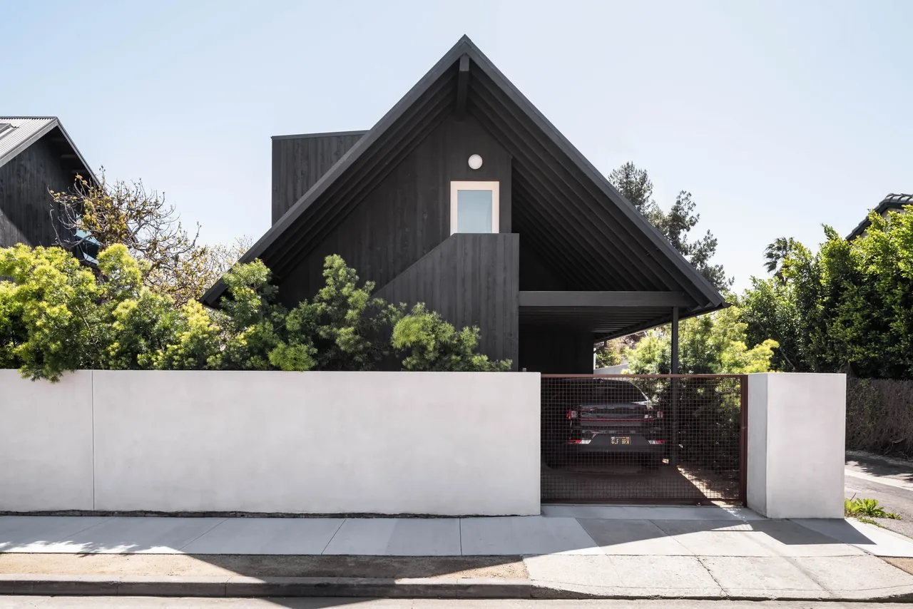 Glencoe Avenue House features a steep gable roof and dark wood siding with a covered carport behind a concrete perimeter wall.