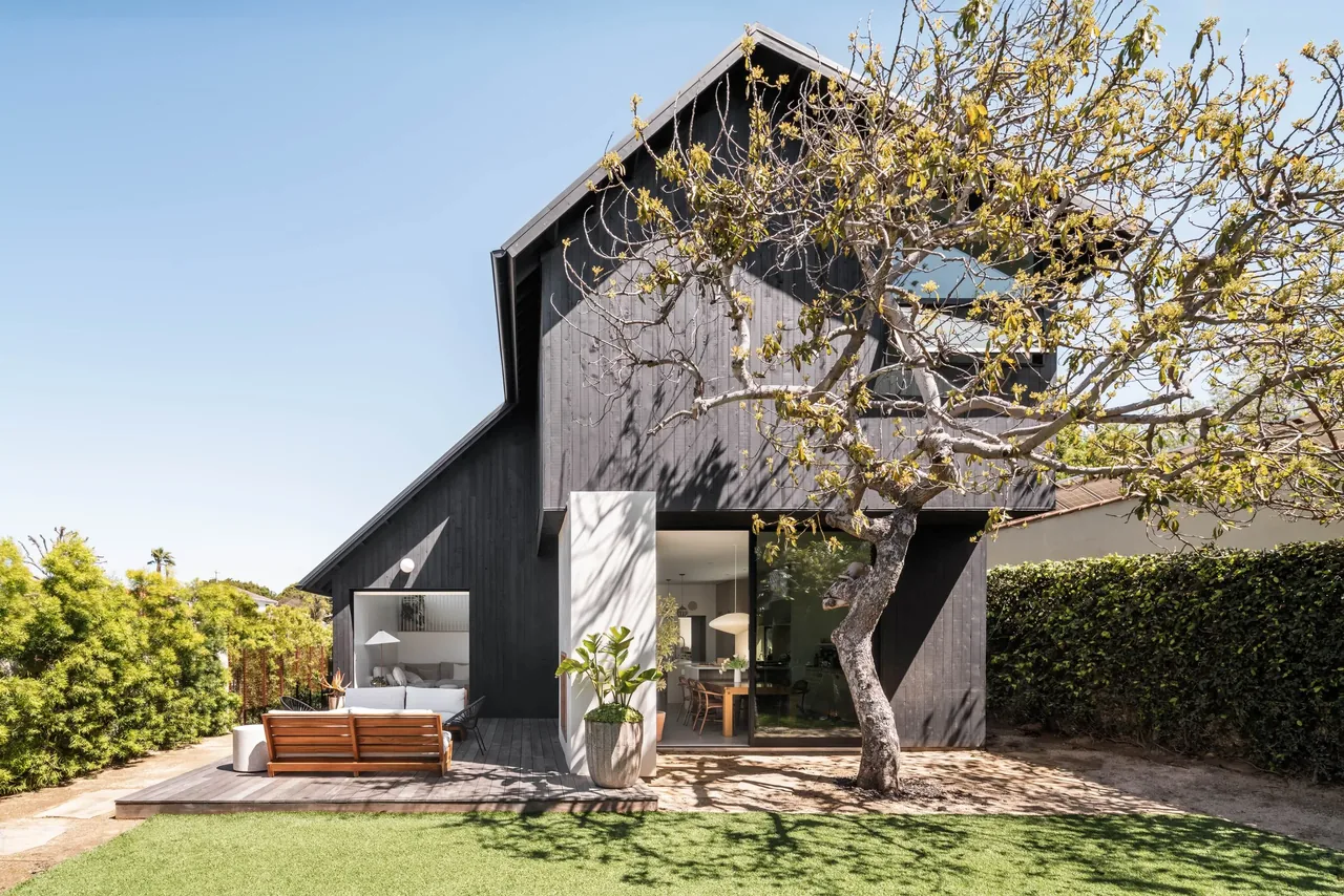 Glencoe Avenue House features dark vertical wood siding with a steep gable roof and an outdoor seating area on a wooden deck.