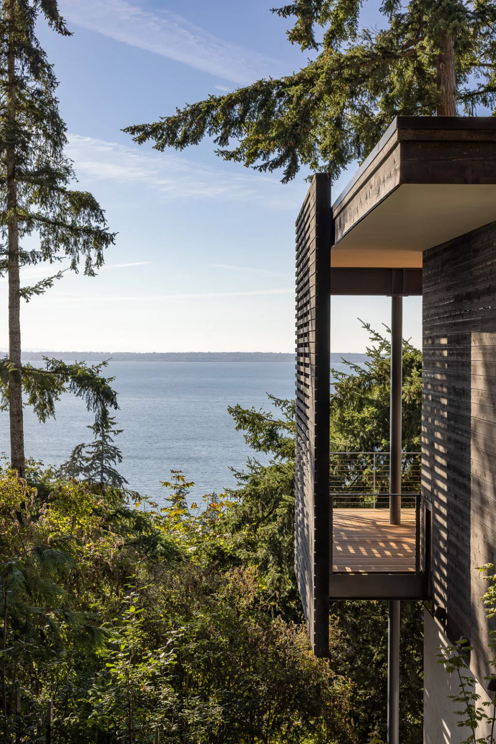 Modern house balcony with horizontal wood slat screen and metal cable railing overlooking water and forest.