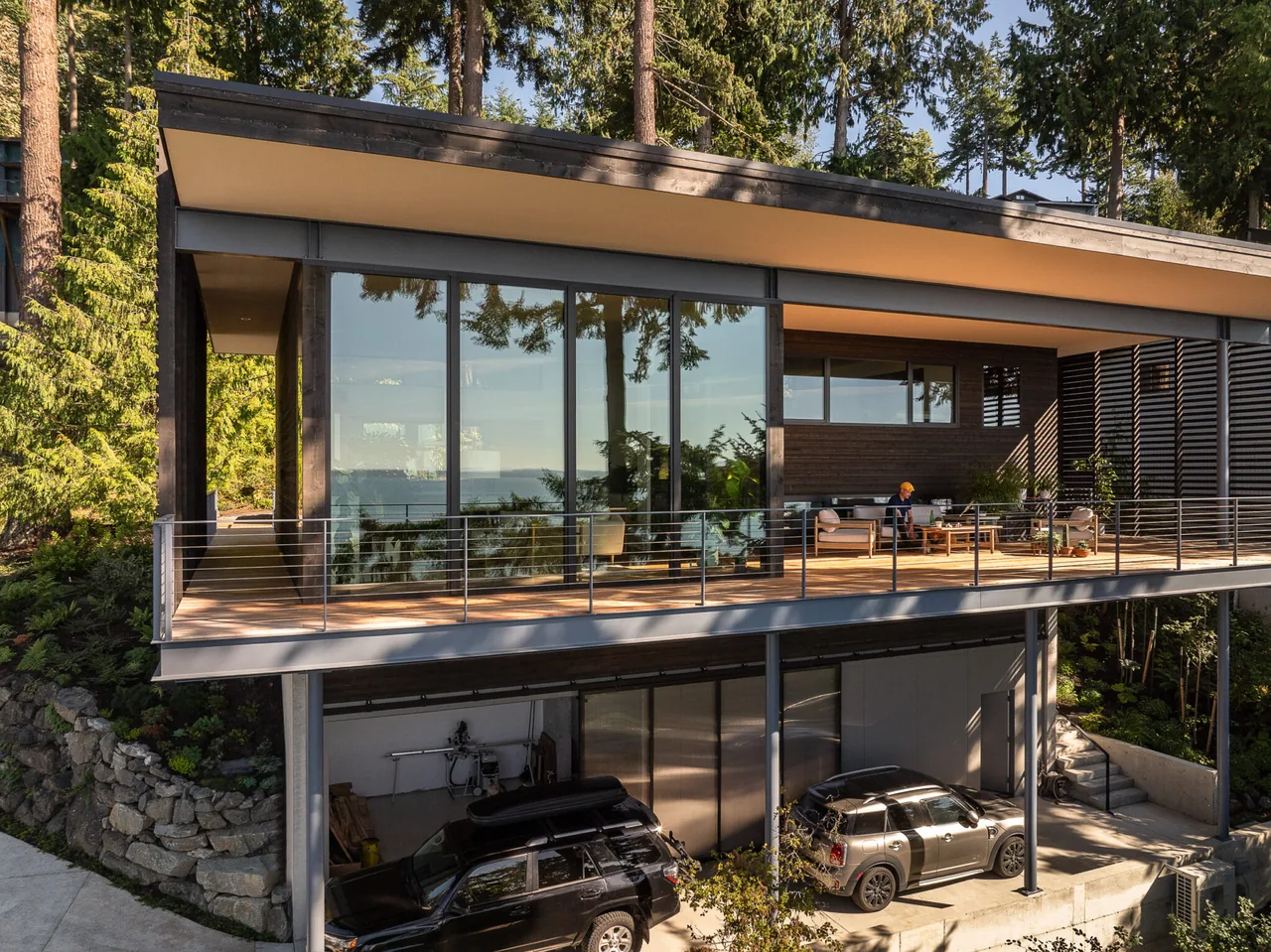 Modern house with an upper-level wooden deck featuring cable railing and large glass windows overlooking a wooded landscape.
