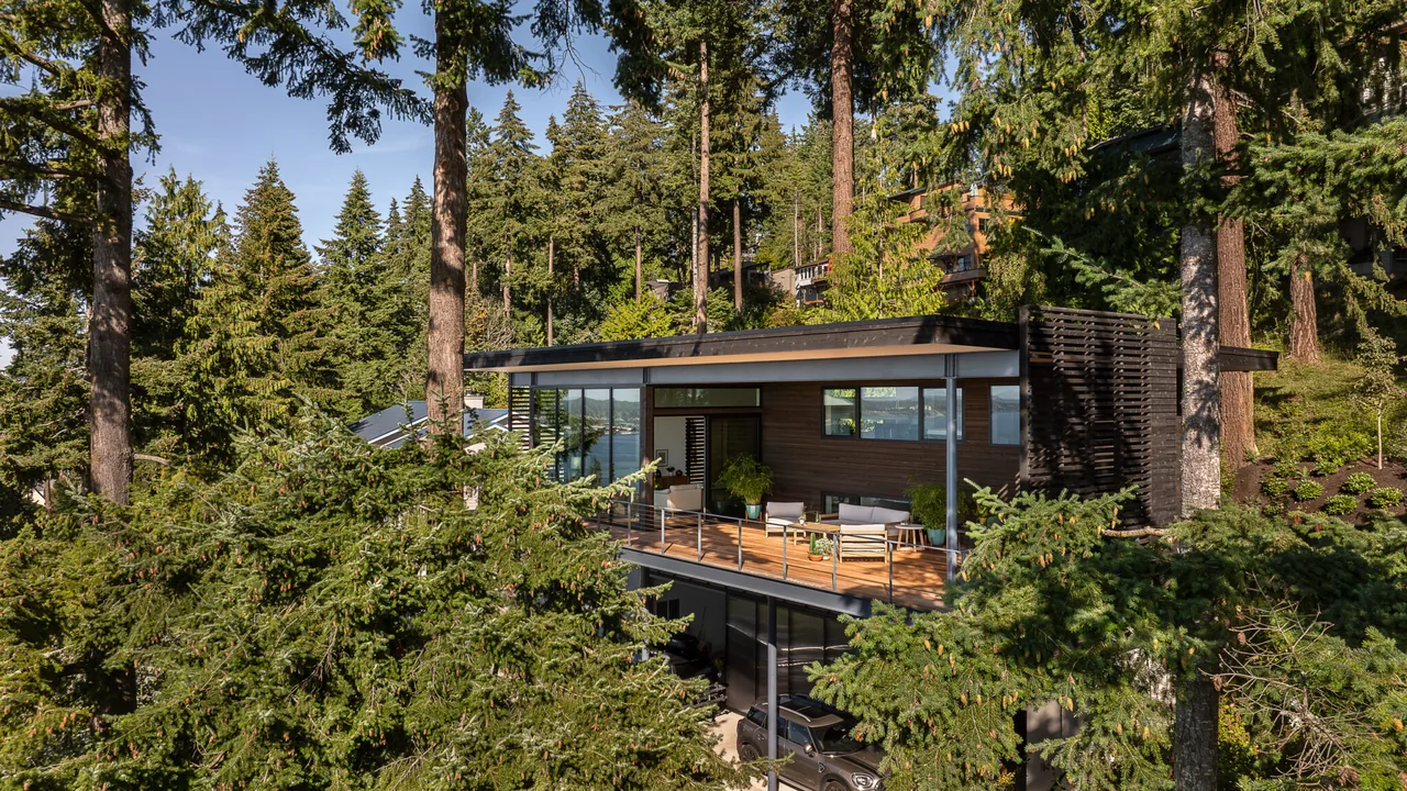 The Nest project features a modern hillside house with a spacious covered deck and cable railing, set amid a dense evergreen forest with water views.