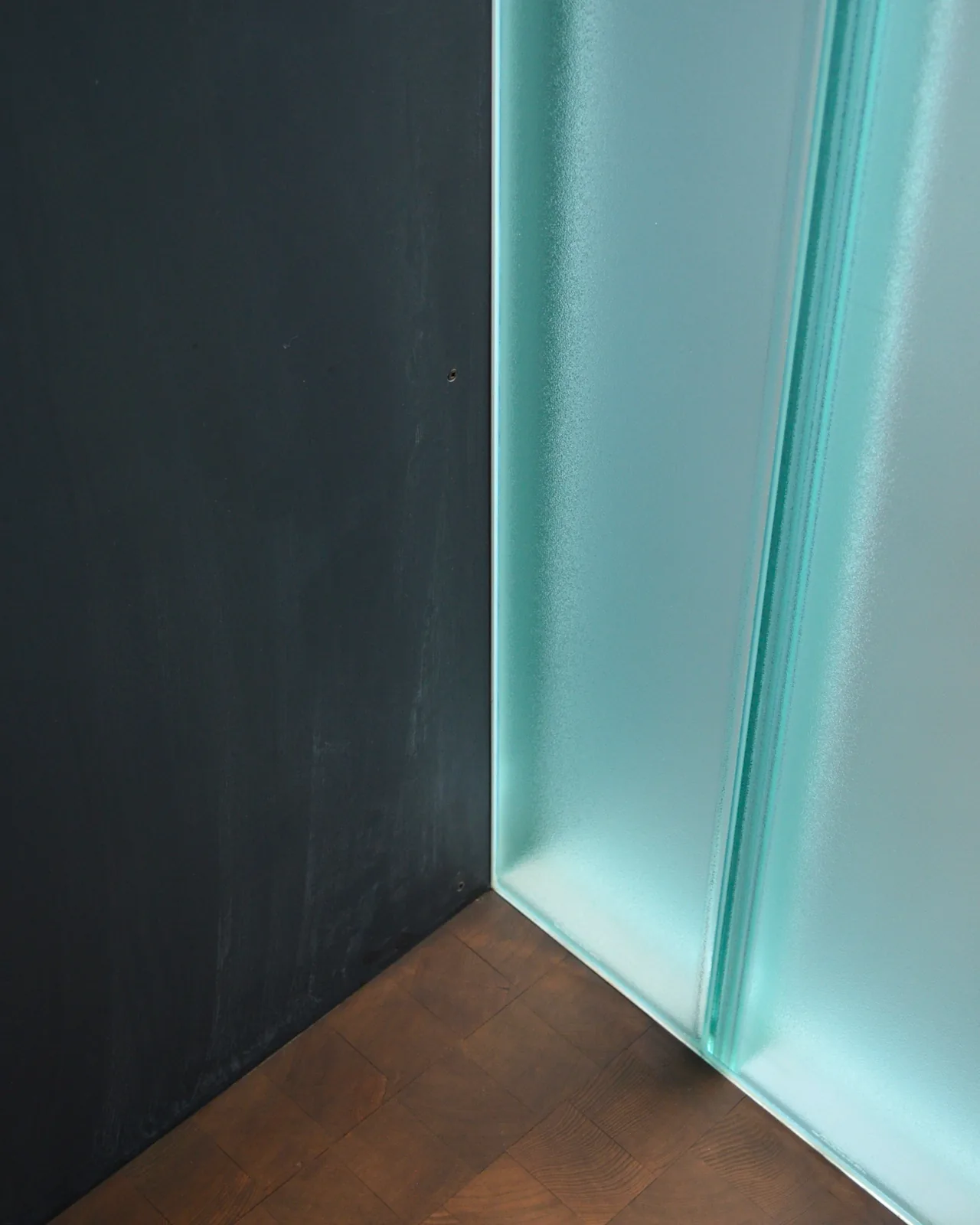 Corner detail of a frosted glass wall next to a dark textured wall with wooden flooring at DMG House.