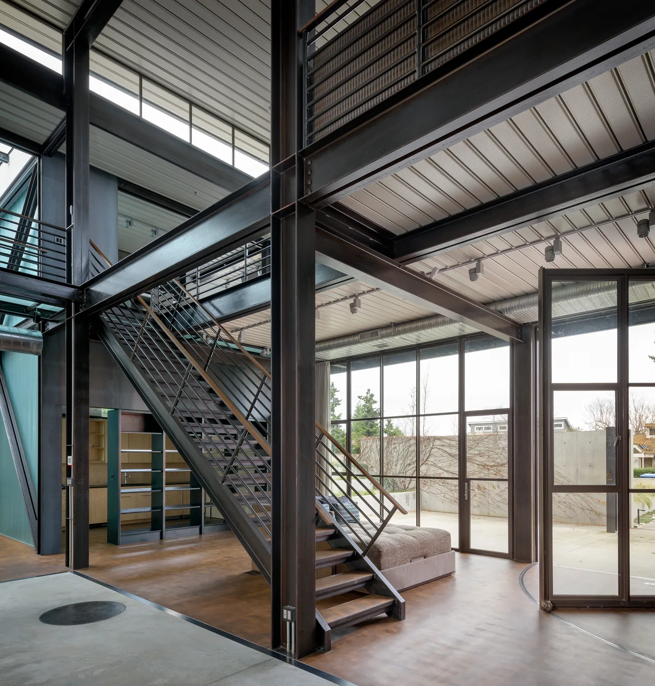 The DMG House interior features exposed black steel framing, a metal stair with railing, and expansive two-story glazing.