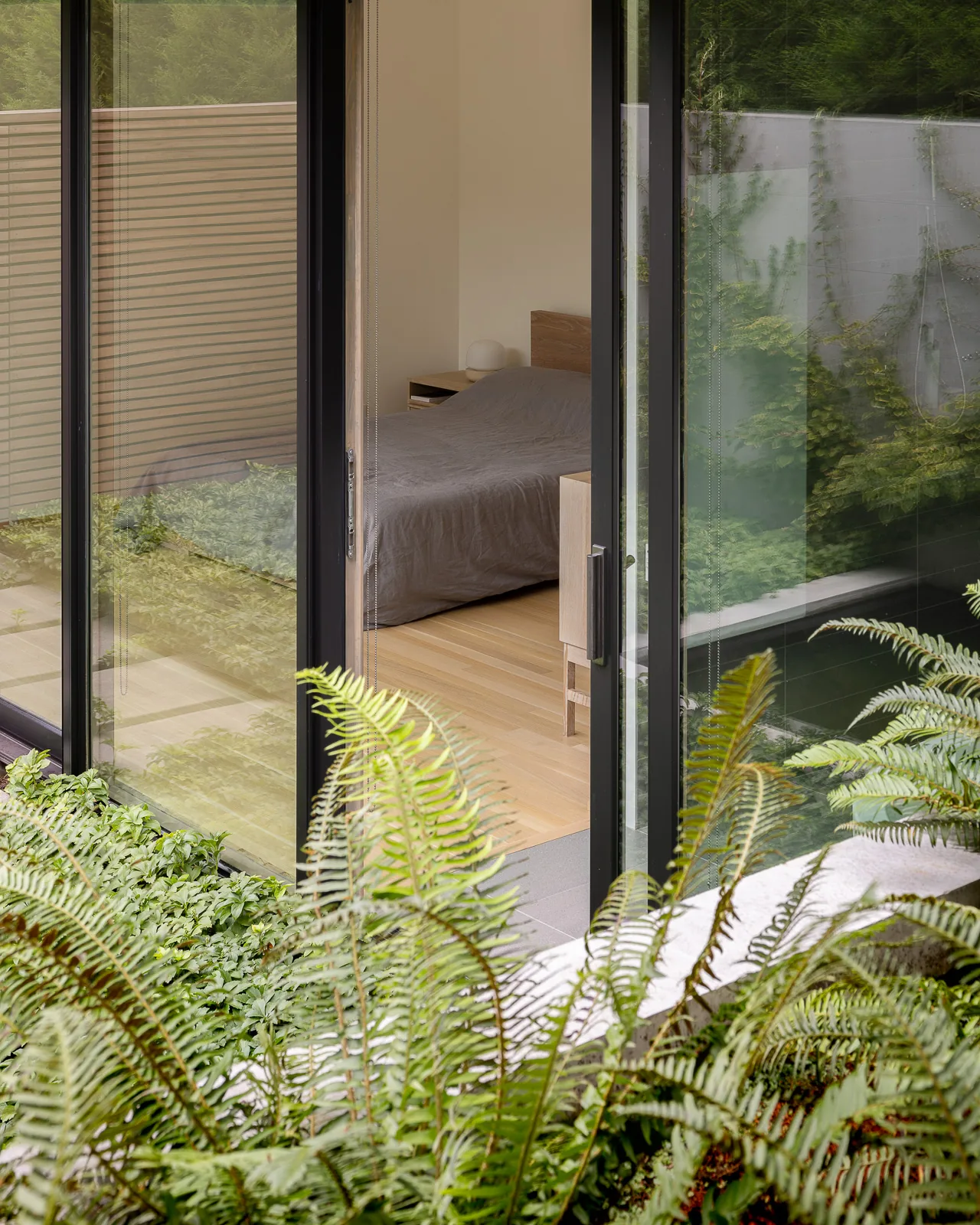 Bedroom with wood floor seen through sliding glass doors with black frames opening to greenery outside.