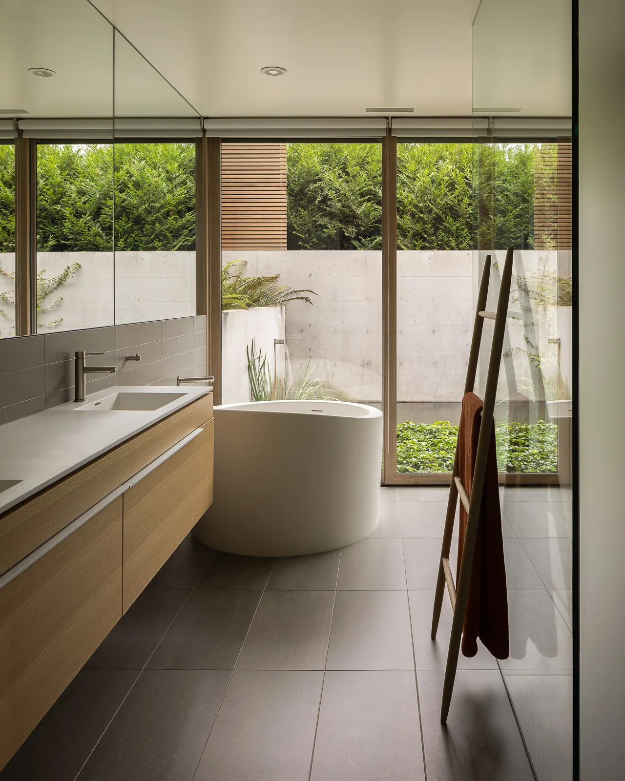 Bathroom with freestanding tub, floating wood vanity, and large windows revealing lush outdoor planting.