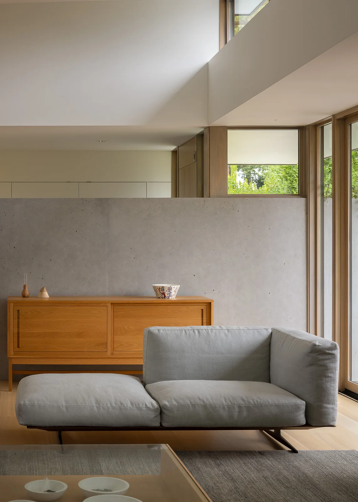 Living area features a minimalist gray sofa, wood sideboard, and concrete accent wall by tall wood-framed windows.