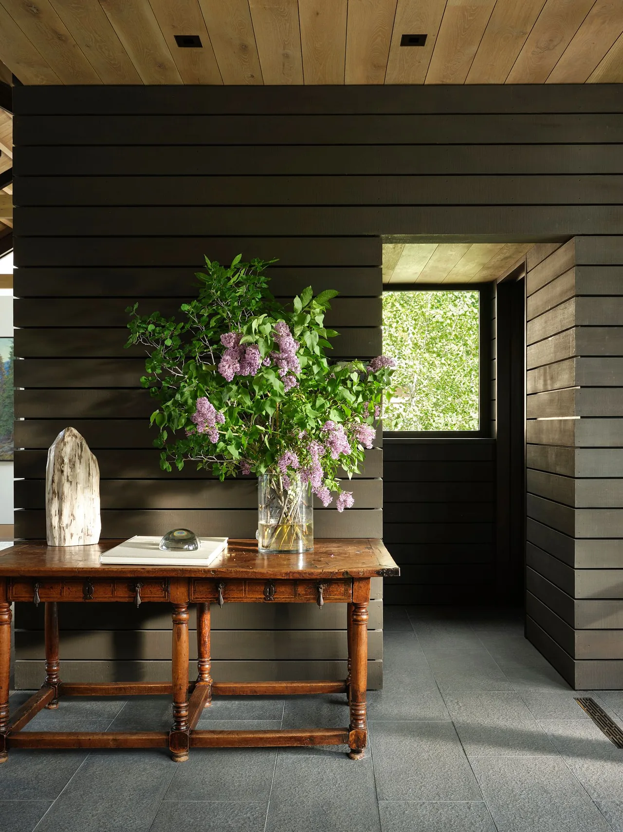 Entry space featuring dark wood horizontal siding, an antique wooden table with lilac flowers, and a view through a square window.