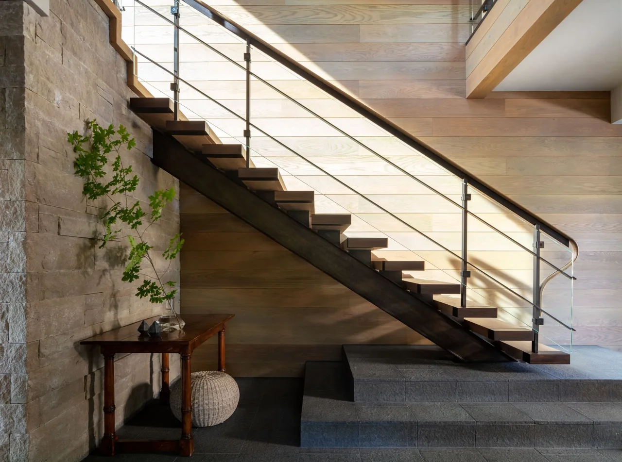 A modern interior staircase features wooden steps with glass and metal railing, set against stone and wood-paneled walls.