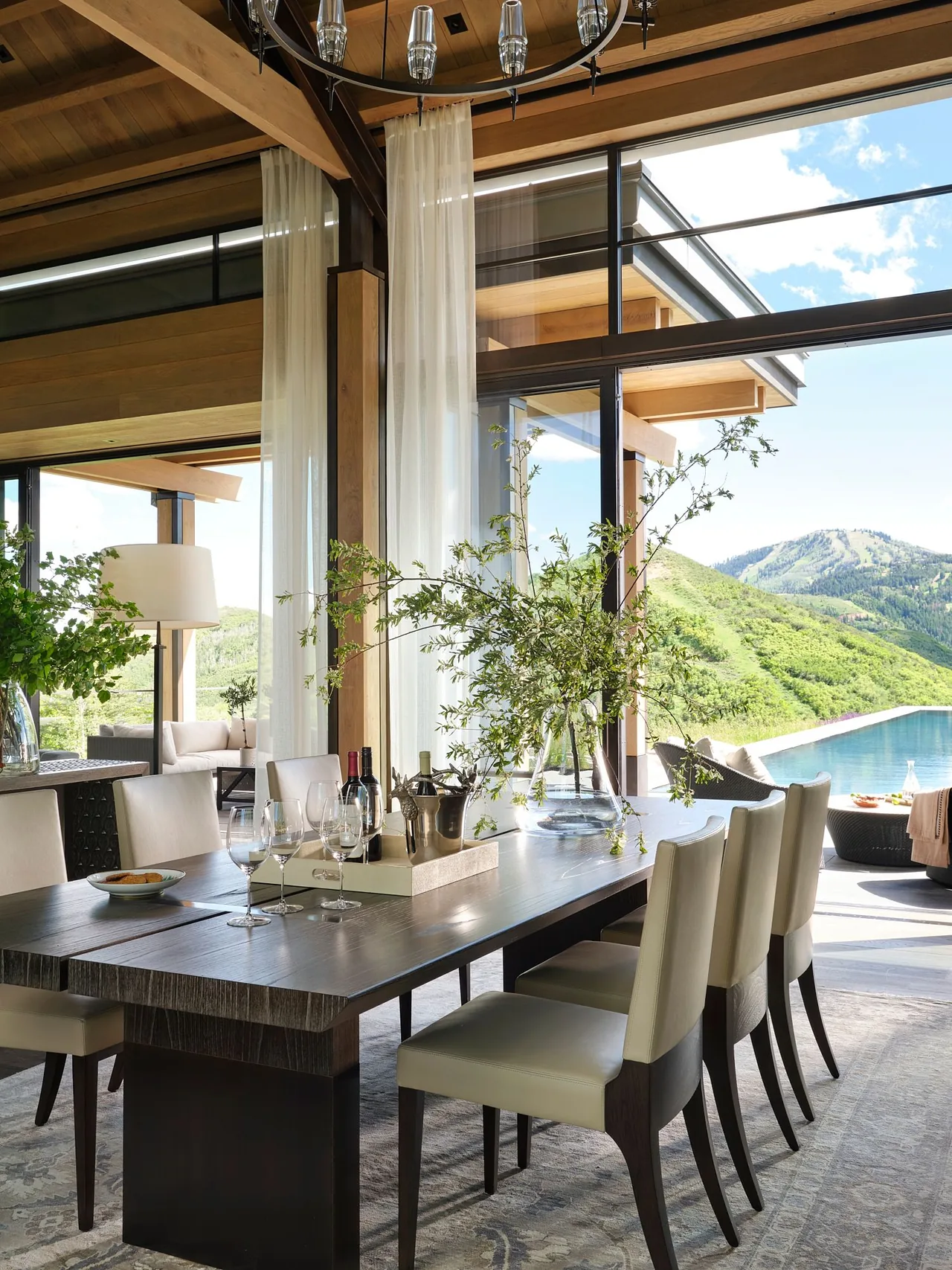 Dining room features a large wood table with cream leather chairs, sheer curtains, and expansive glass windows overlooking the infinity pool and mountains.