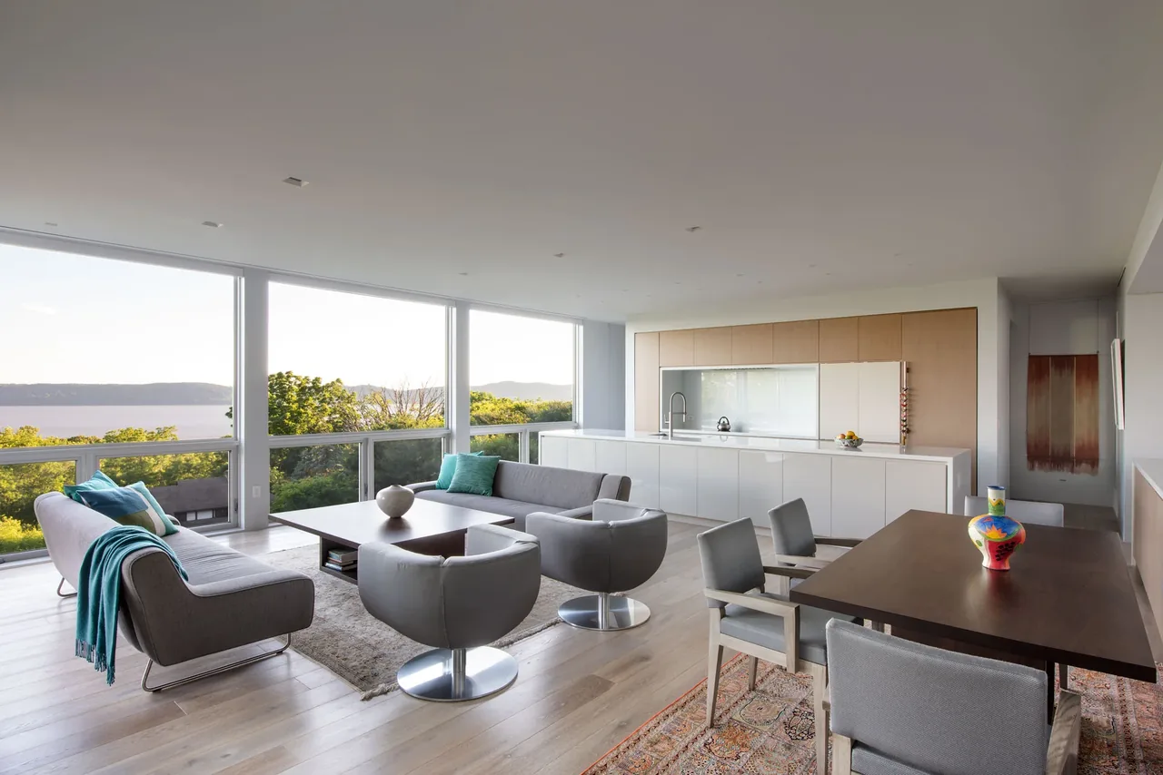 A modern open living, dining, and kitchen space with floor-to-ceiling windows overlooking a river view.