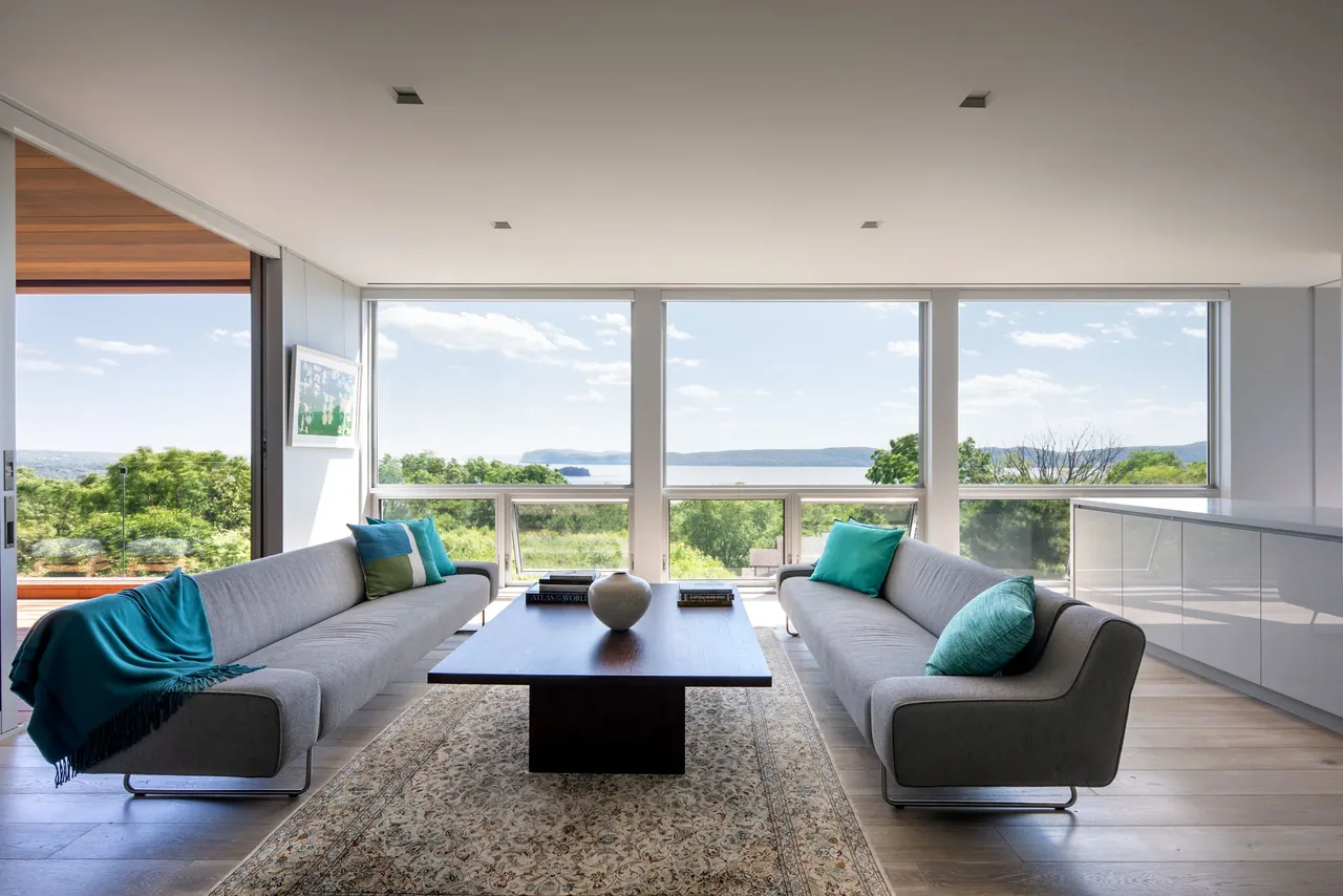 Living room of Hudson River House with floor-to-ceiling glazing offering river views, furnished with gray sofas and a wood coffee table.