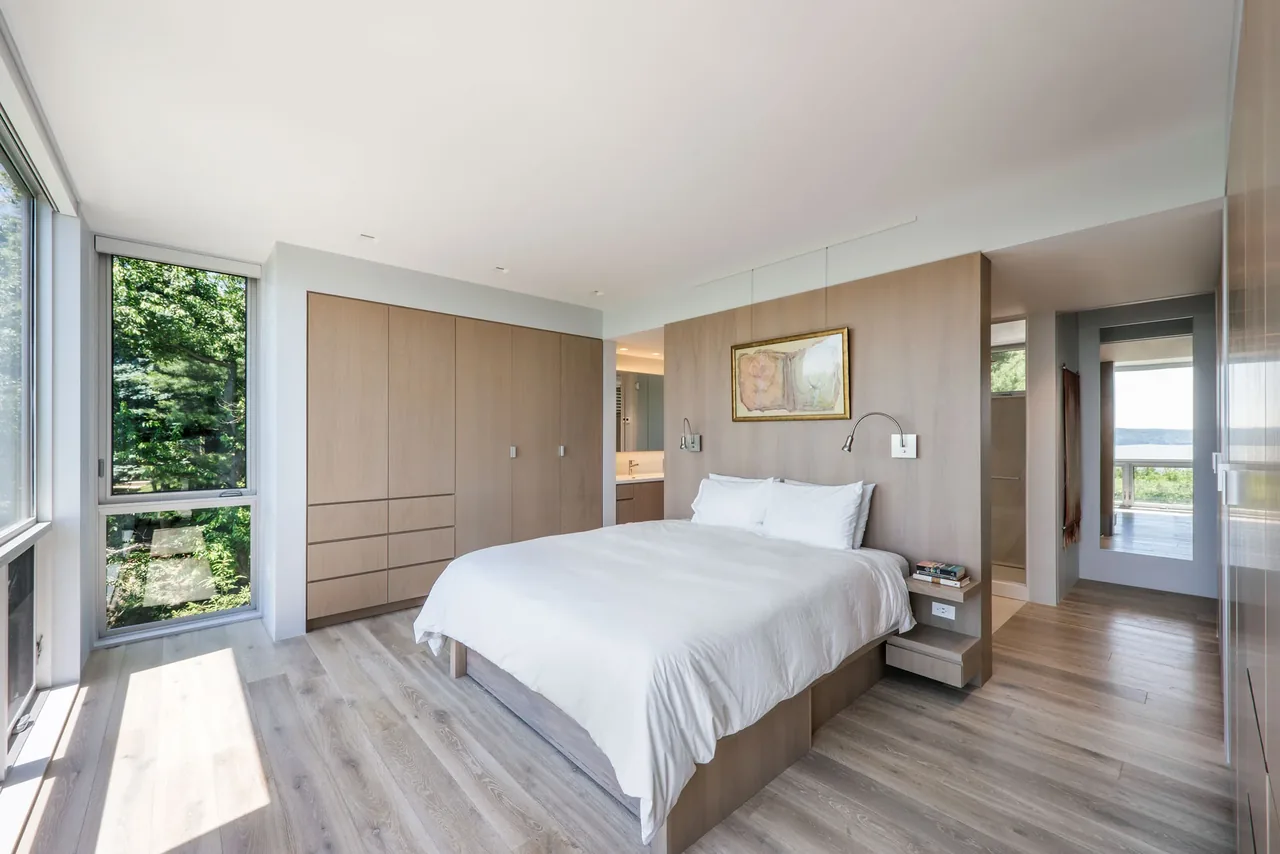 Modern bedroom with floor-to-ceiling windows and wood cabinetry, overlooking the river through an open hallway.