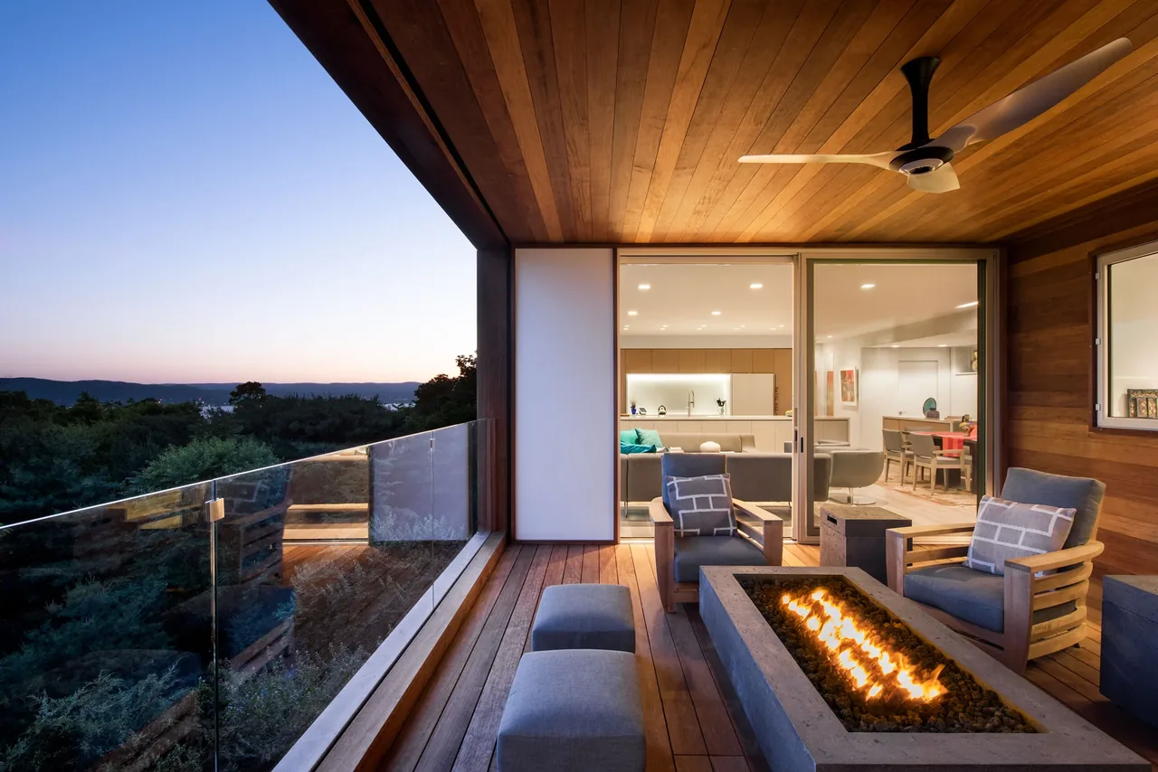 Covered deck with modern outdoor seating and fire pit looks out over a tree-lined view at dusk.