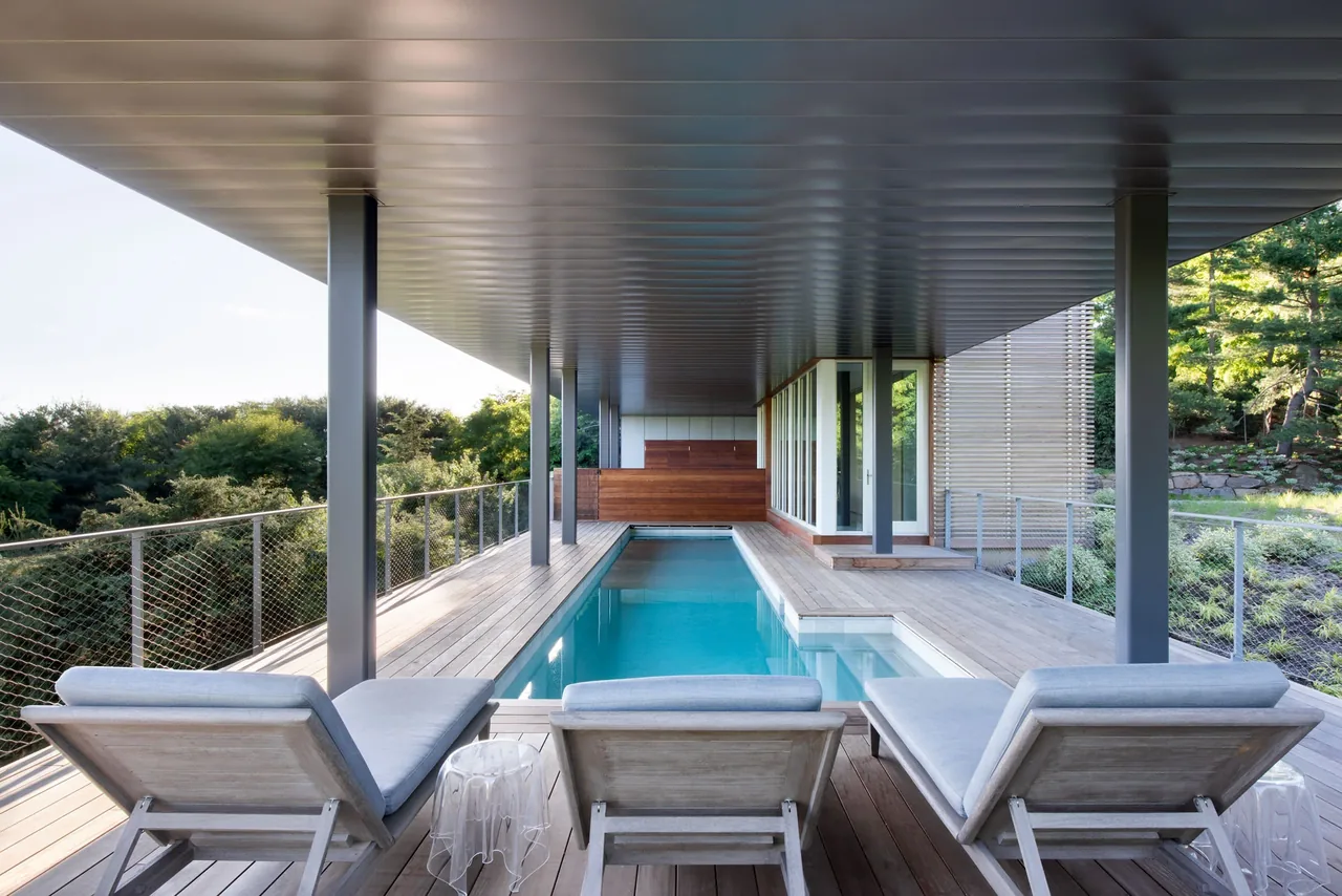 Covered outdoor pool area with wooden deck and three lounge chairs overlooking greenery.