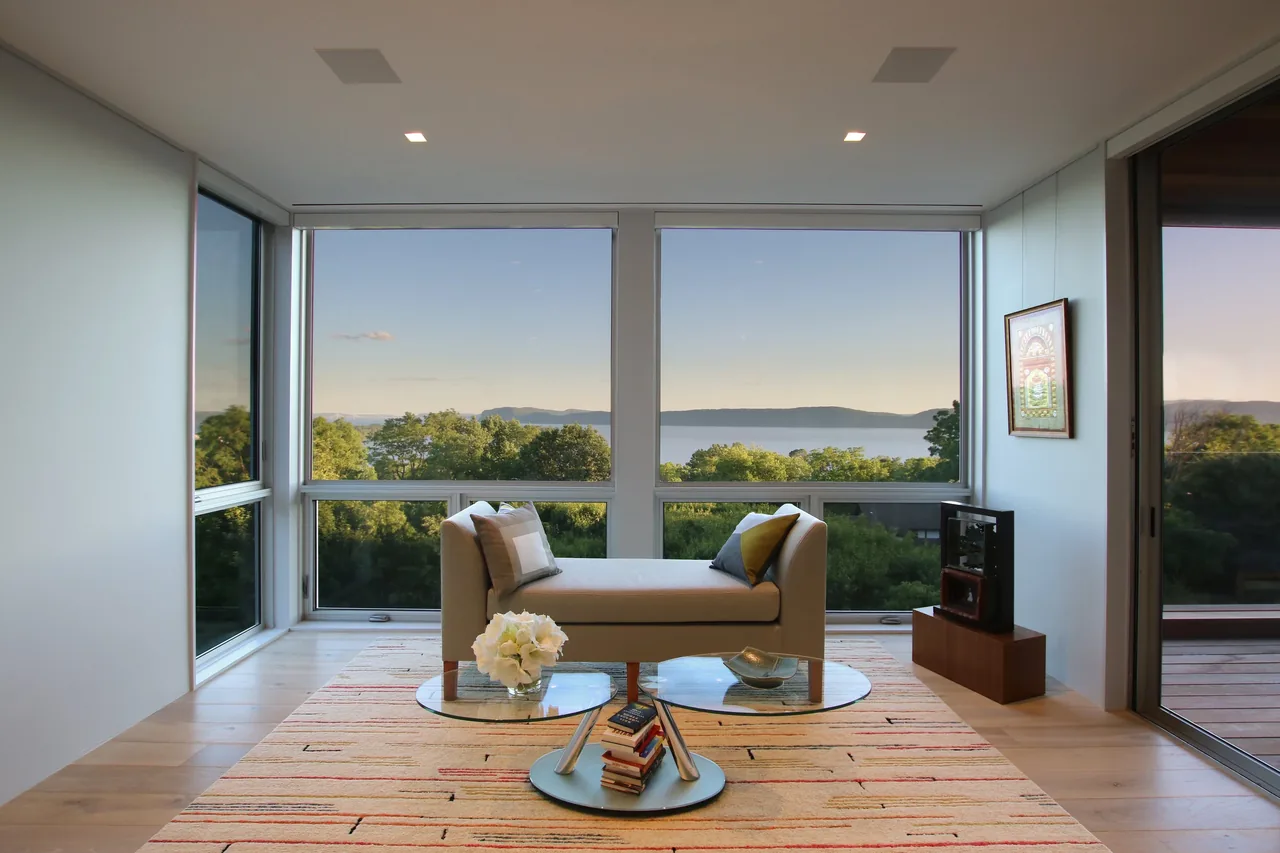 Living room with floor-to-ceiling corner windows overlooking river and trees, furnished with a modern chaise lounge and glass coffee tables.