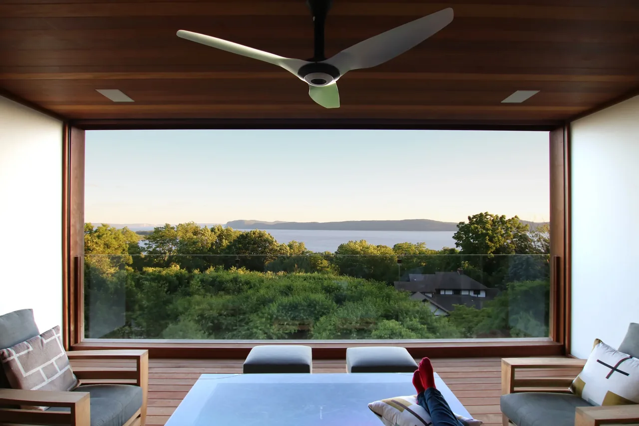 Outdoor living space framed by wood ceiling with ceiling fan and glass railing showing river and tree landscape view.