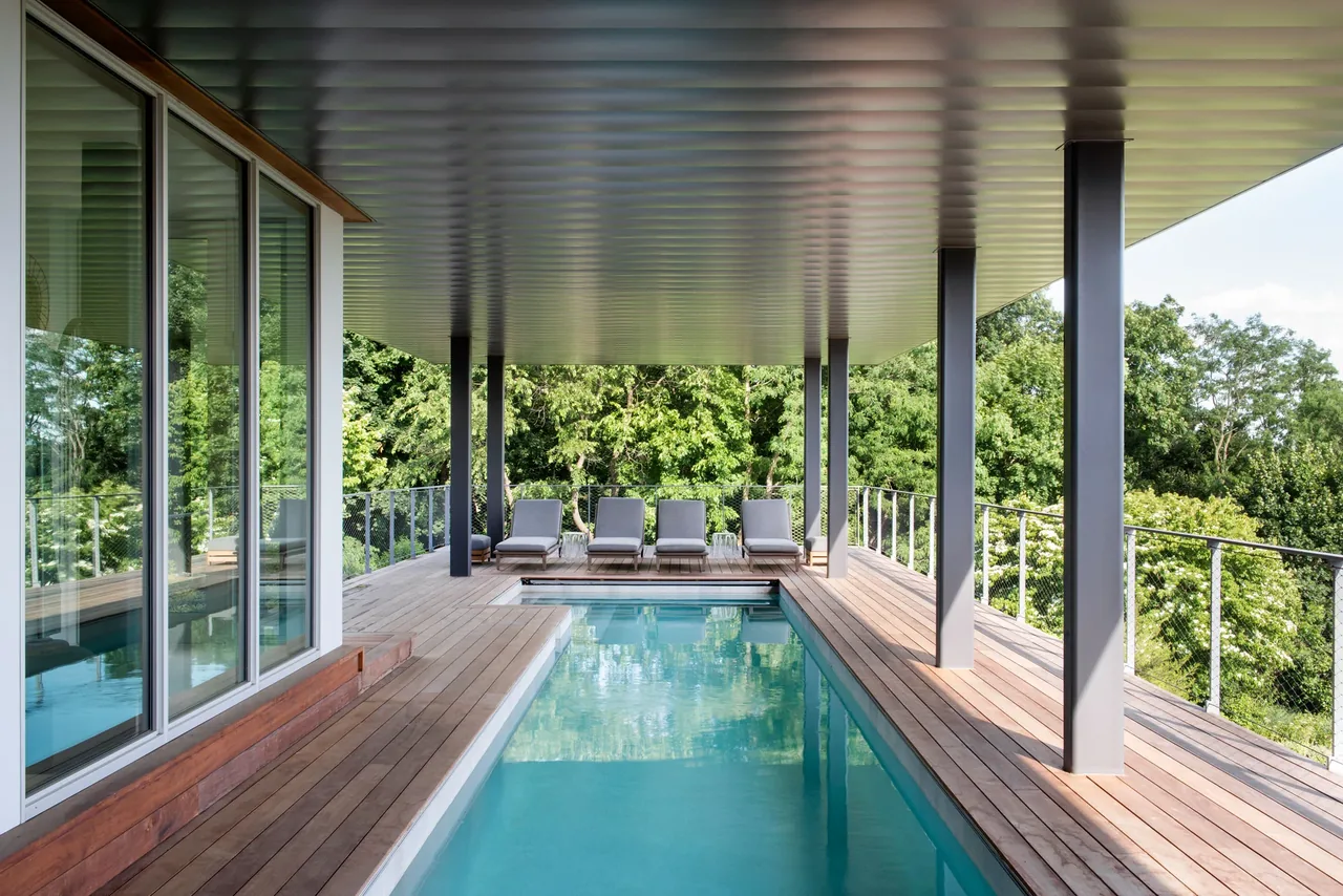 Covered outdoor living space features a rectangular pool with wooden decking and seating at the far end.