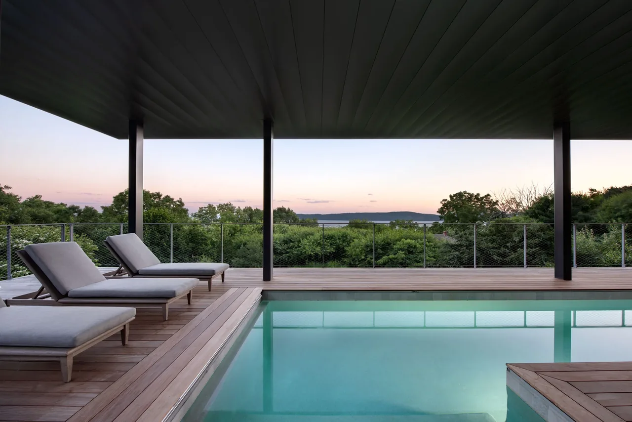Pool deck with seating and covered roof offers a river view and scenic greenery in Hudson Valley, NY.