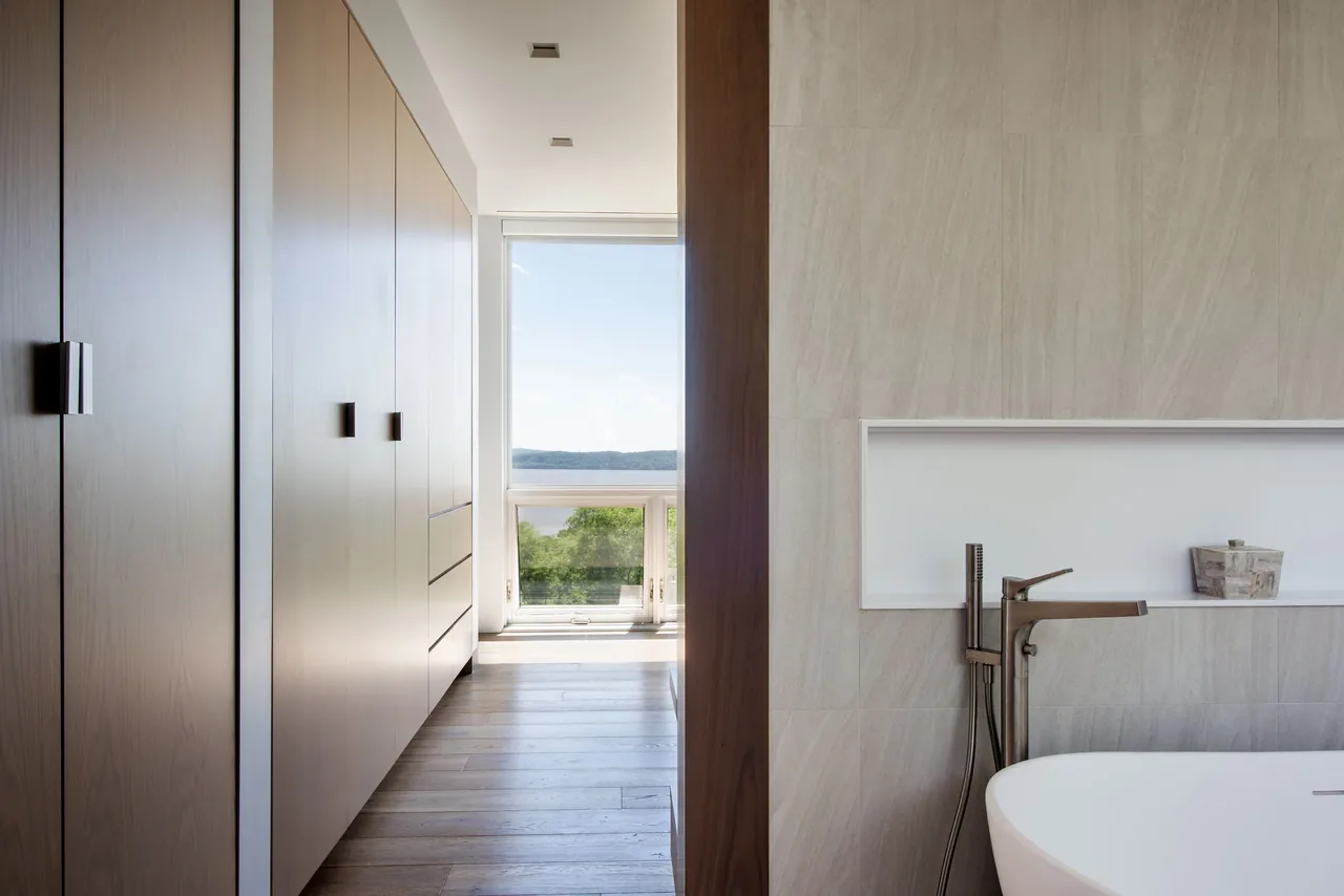 Hallway with built-in wooden cabinetry leads to a large window with a river view beside a freestanding tub.
