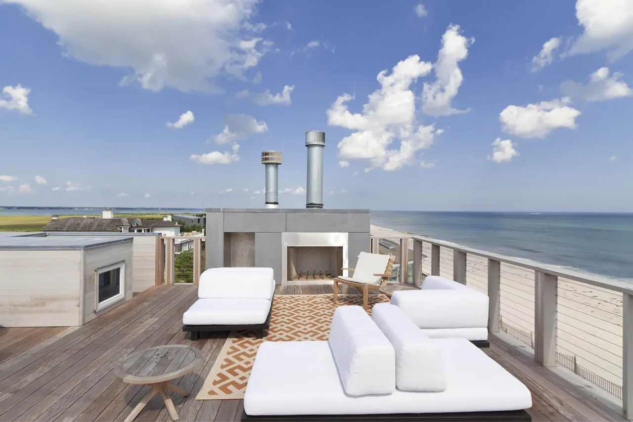 Outdoor living space on a wooden deck with modern white seating and a built-in fireplace facing an ocean view at the Dune Road Beach House.