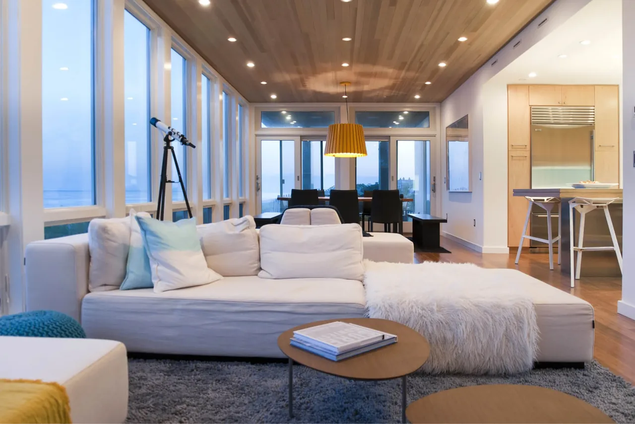 Living room with a beige sectional sofa and a dining area showcasing wide windows overlooking the ocean at Dune Road Beach House.