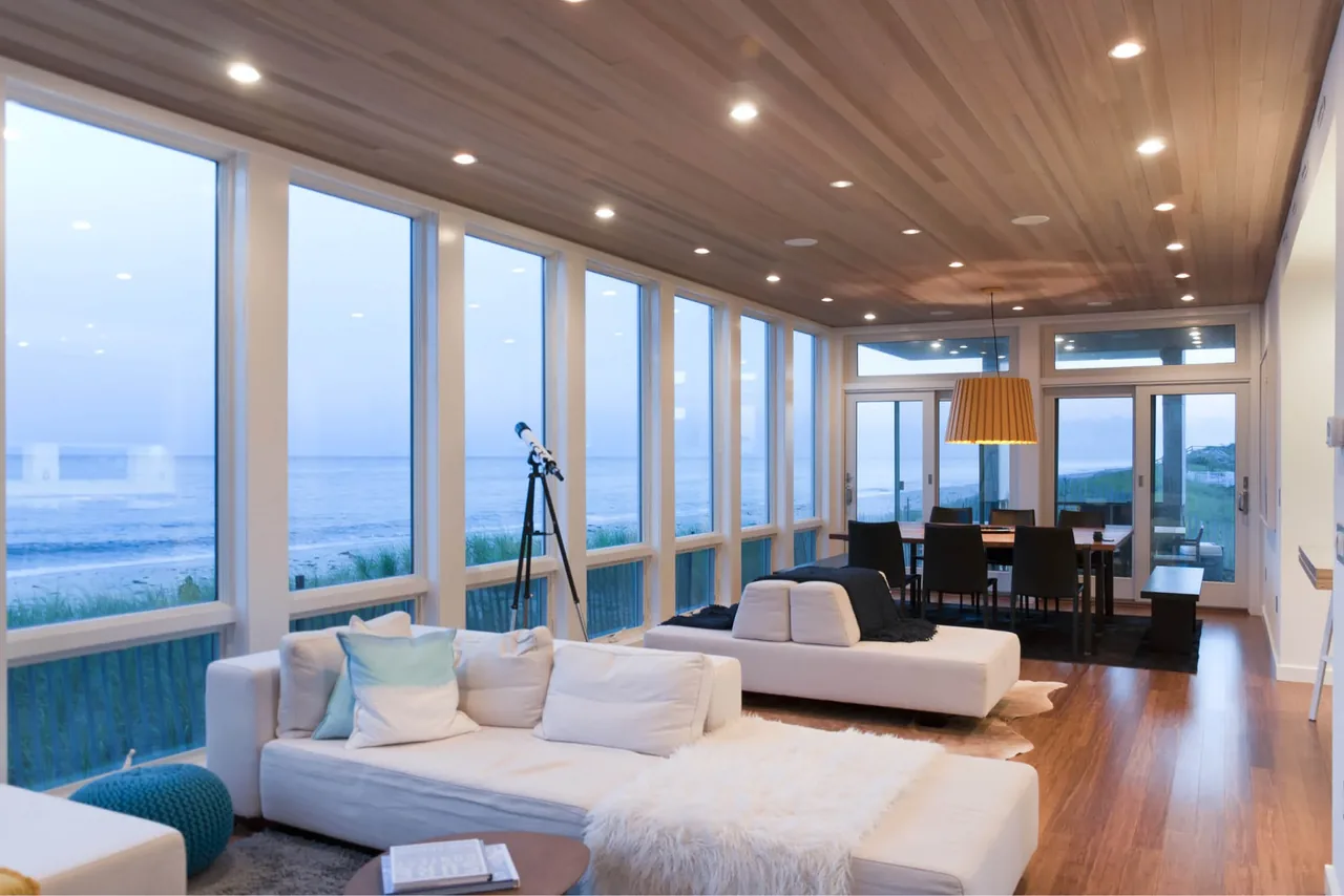 Open living and dining space with wood ceiling and floor-to-ceiling windows overlooking the ocean at Dune Road Beach House.
