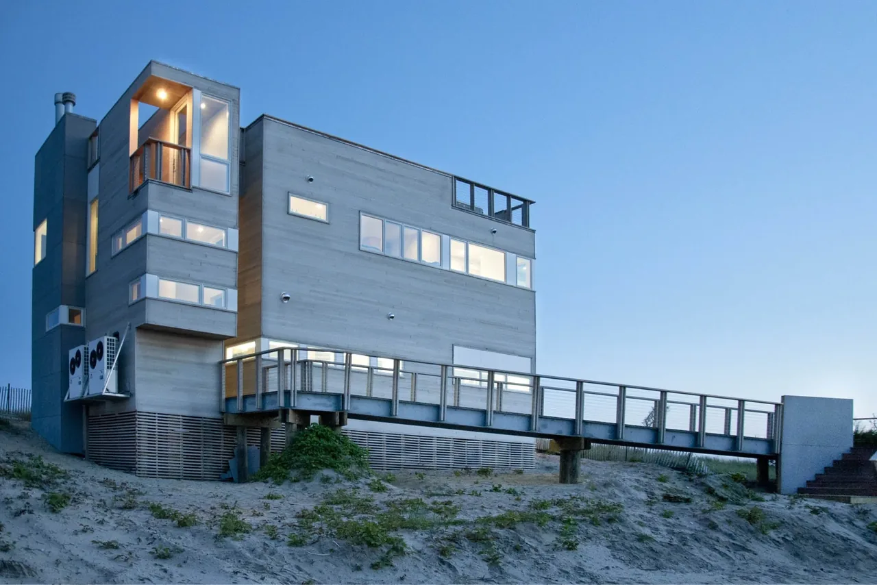 Dune Road Beach House in East Quogue, NY, features wide horizontal windows and an elevated deck with railing.