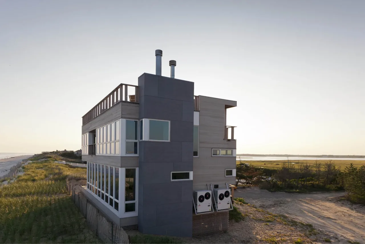 Dune Road Beach House in East Quogue, NY features wide horizontal windows and a rooftop deck overlooking coastal dunes.