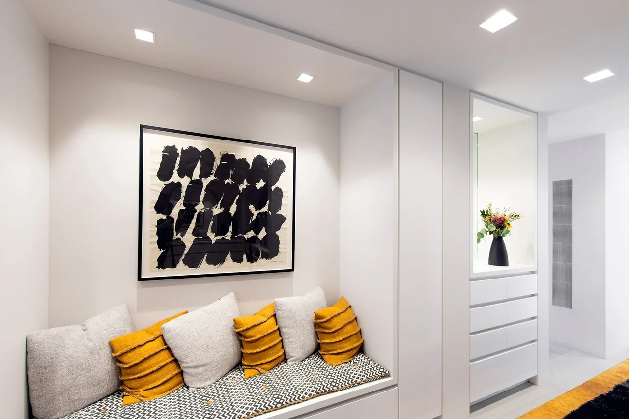 Seating nook with patterned cushion, orange and white pillows, and abstract art above in a bright minimalist interior.