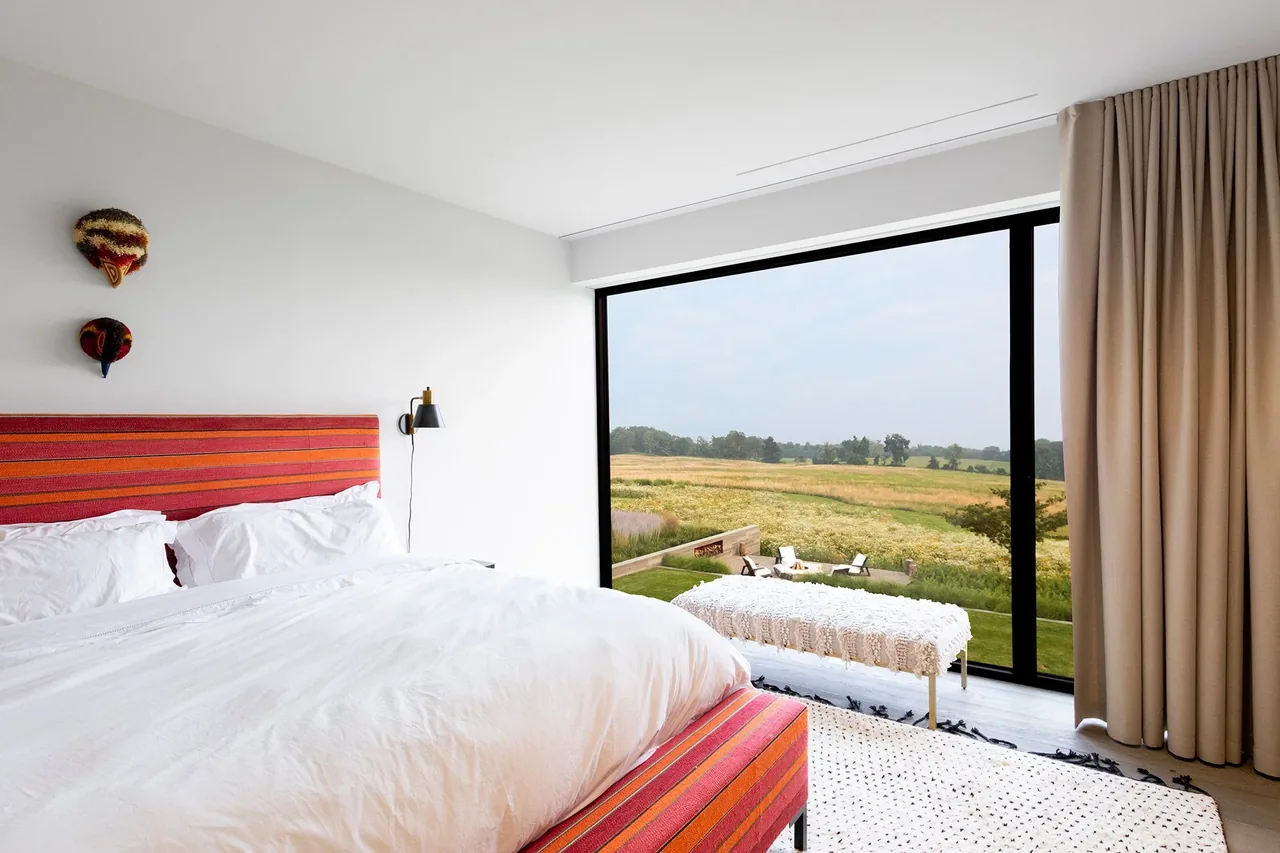 Bedroom in the Millbrook Residence with expansive window overlooking rural landscape, featuring red-striped bed upholstery.