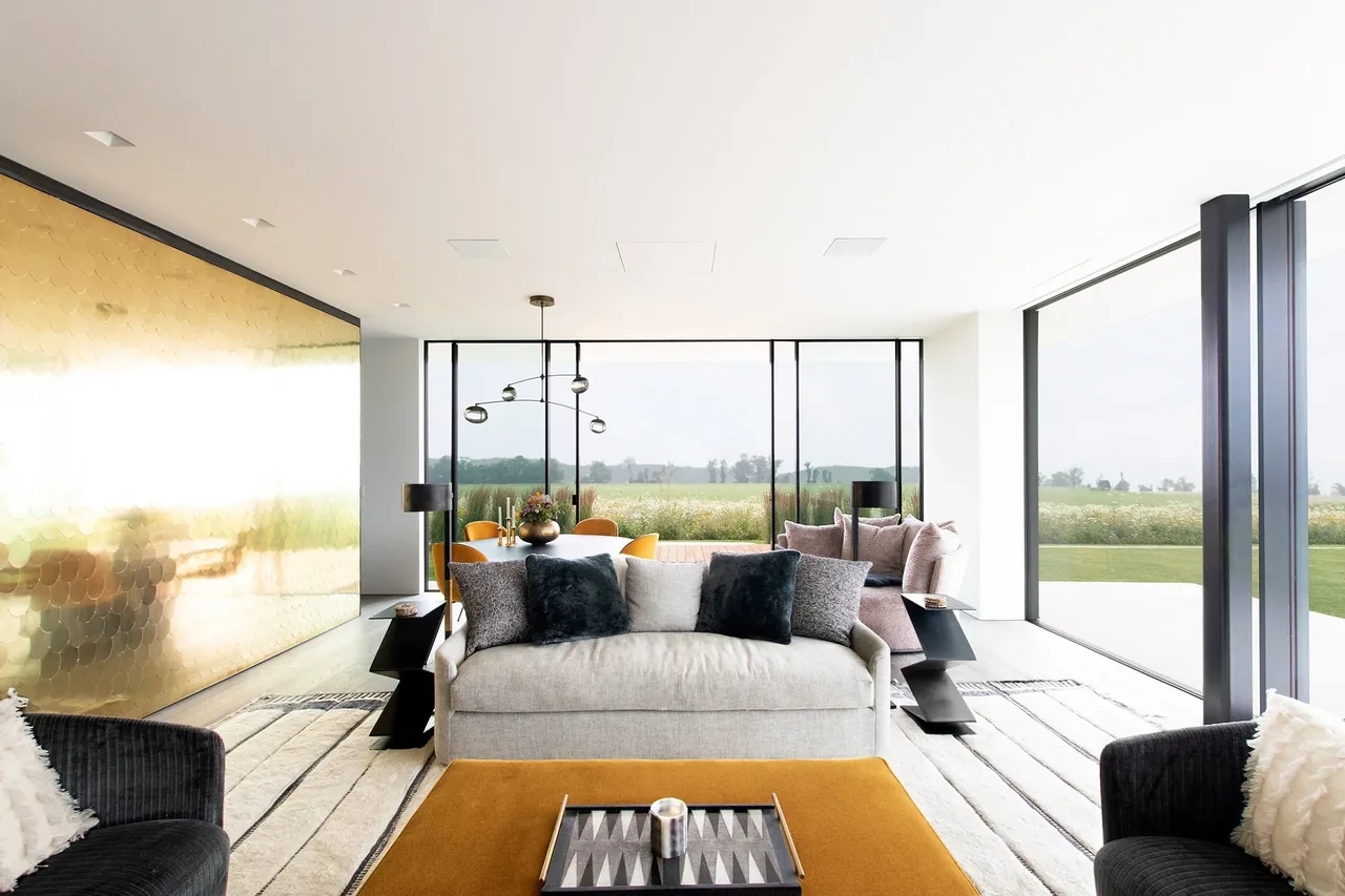 Living room with a large beige sofa, black pillows, and floor-to-ceiling glazing showing a view of a green meadow.