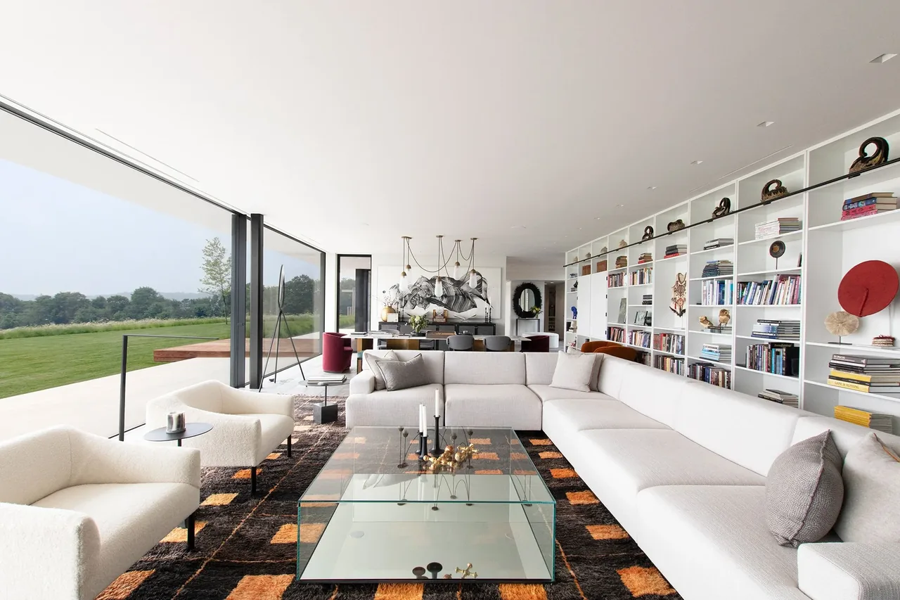 Millbrook Residence living room features floor-to-ceiling glazing with views of the green meadow and built-in white bookshelf wall.