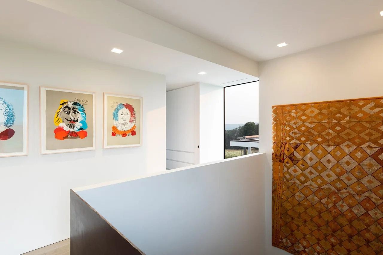 Stair landing with three framed artworks and a large textile hanging beside a window overlooking the landscape.