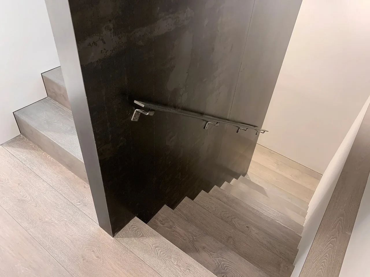 Wooden staircase with light gray steps next to a black wall panel featuring a metal handrail in the Millbrook Residence.