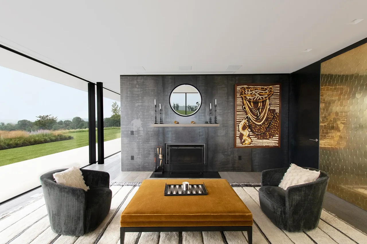 The Millbrook Residence living room features black velvet chairs, a mustard ottoman, and a fireplace with a round mirror above it.
