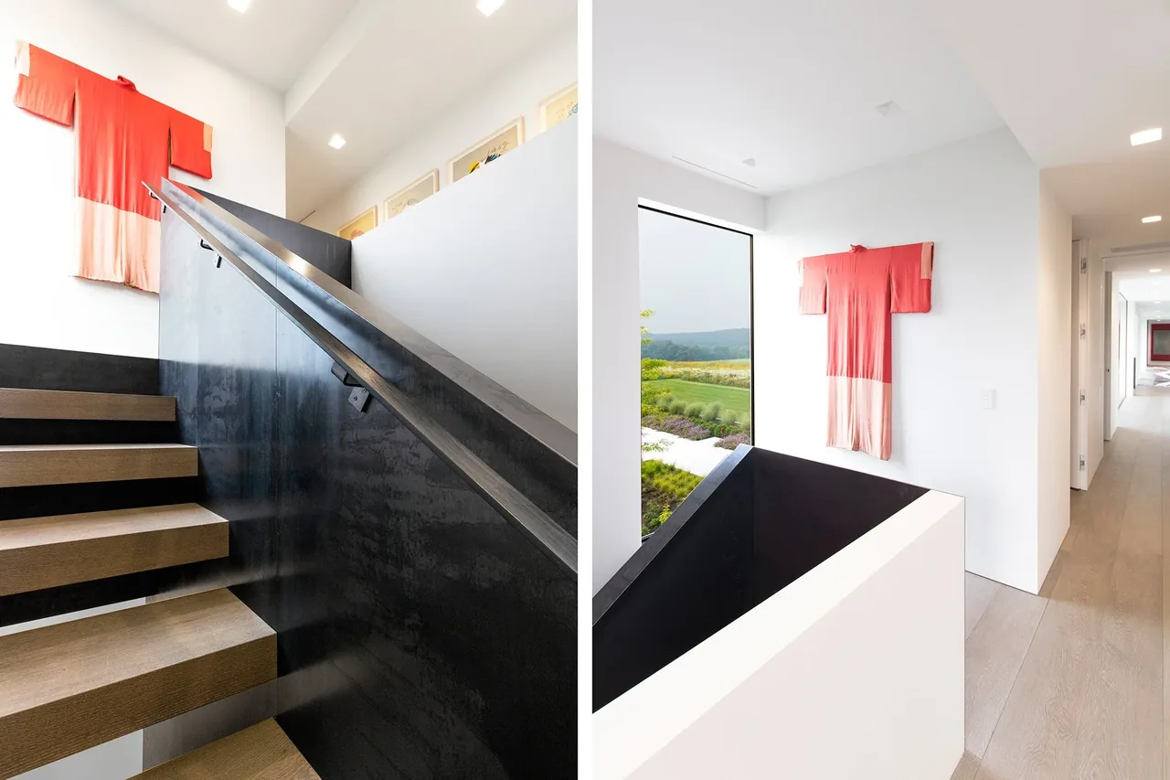 Millbrook Residence stairway features floating wood steps with a black metal handrail beside a white wall adorned with fabric art and a landscape view window.