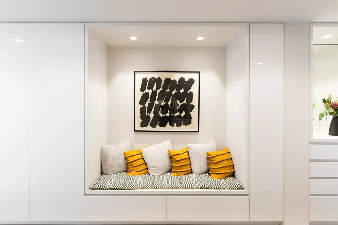 Built-in bench seat with patterned cushion and colorful pillows under abstract artwork in a white room.