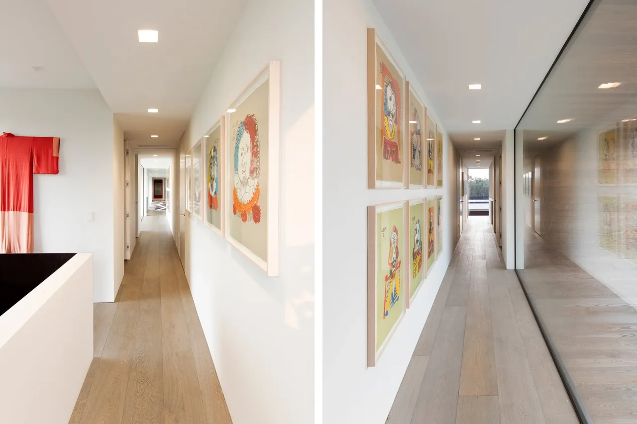 Minimalist hallway with light wood floors, recessed lighting, and framed artwork on white walls, featuring a large reflective glass panel on one side.