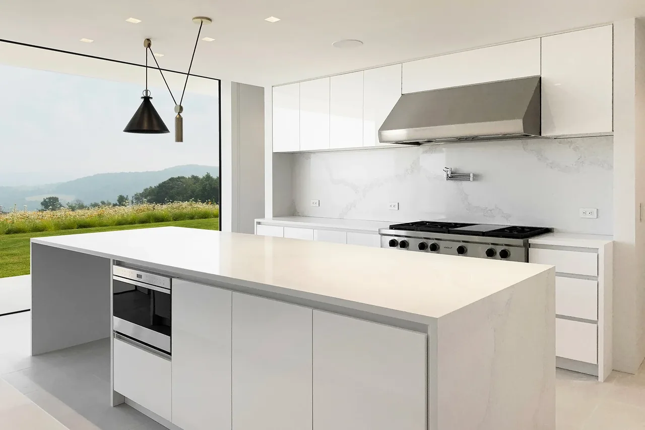 Kitchen with white cabinetry, large island, black cone pendant light, stainless steel range hood, and gas cooktop overlooking a green landscape.