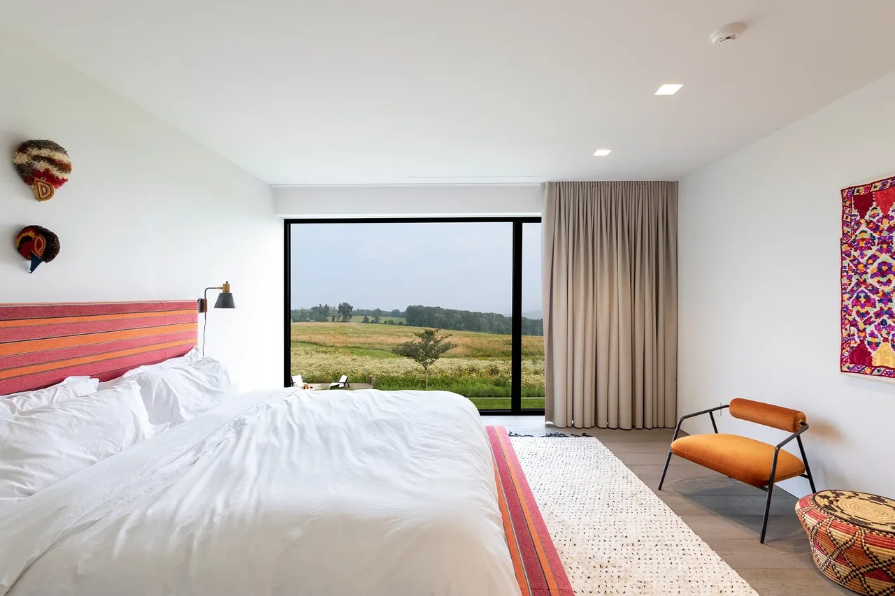 The Millbrook Residence bedroom features a large black-framed window overlooking fields, a striped headboard, and an orange accent chair.