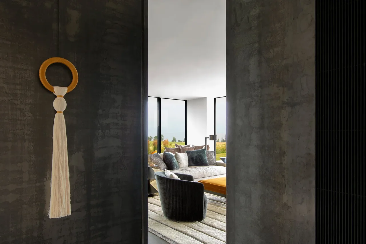Living room with white sectional sofa, large windows showing green meadow, and a hanging textile wall art on dark textured wall.
